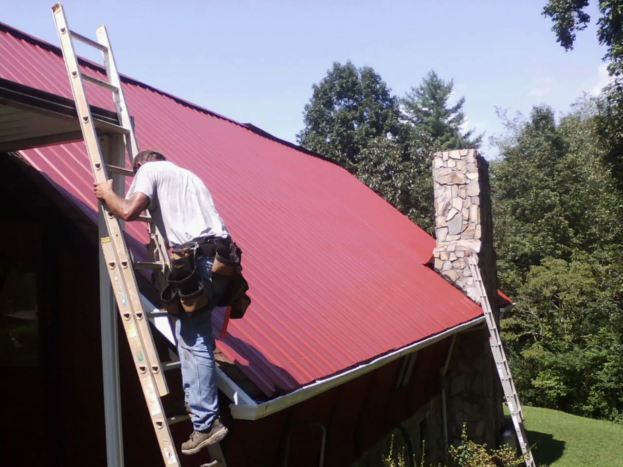 Roofer on a ladder beside a red metal roof and stone chimney — Soffit & Fascia Repair by Roofing Brothers in North Brookfield